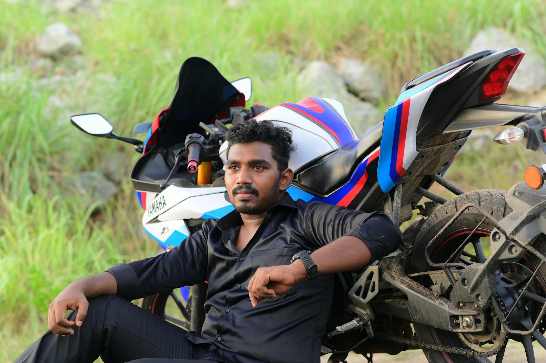 Portrait of a man in a black shirt sitting beside a colorful motorbike in a rural area.