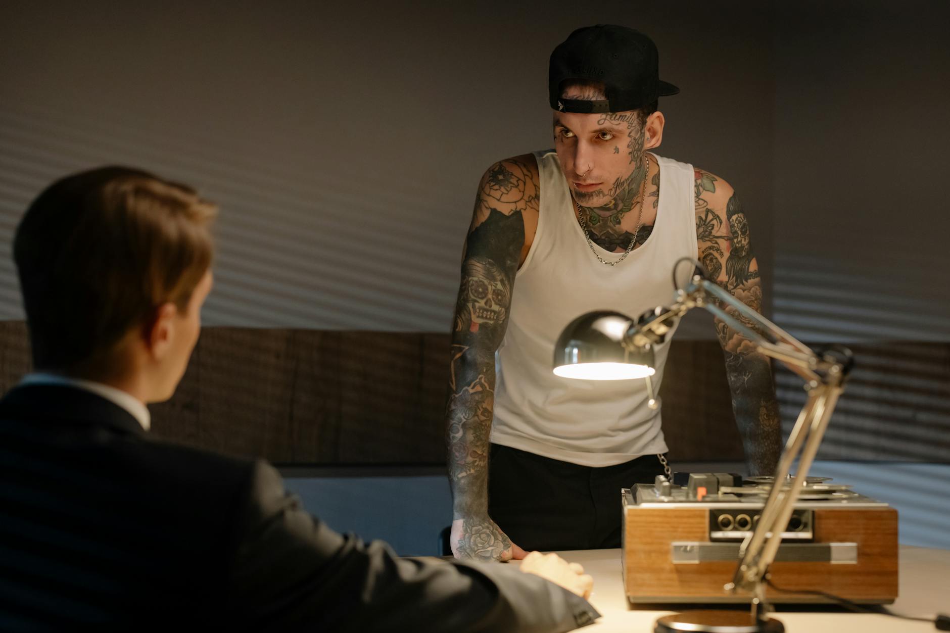 Tattooed man in a police interrogation room leaning over a desk with a tape recorder and lamp.