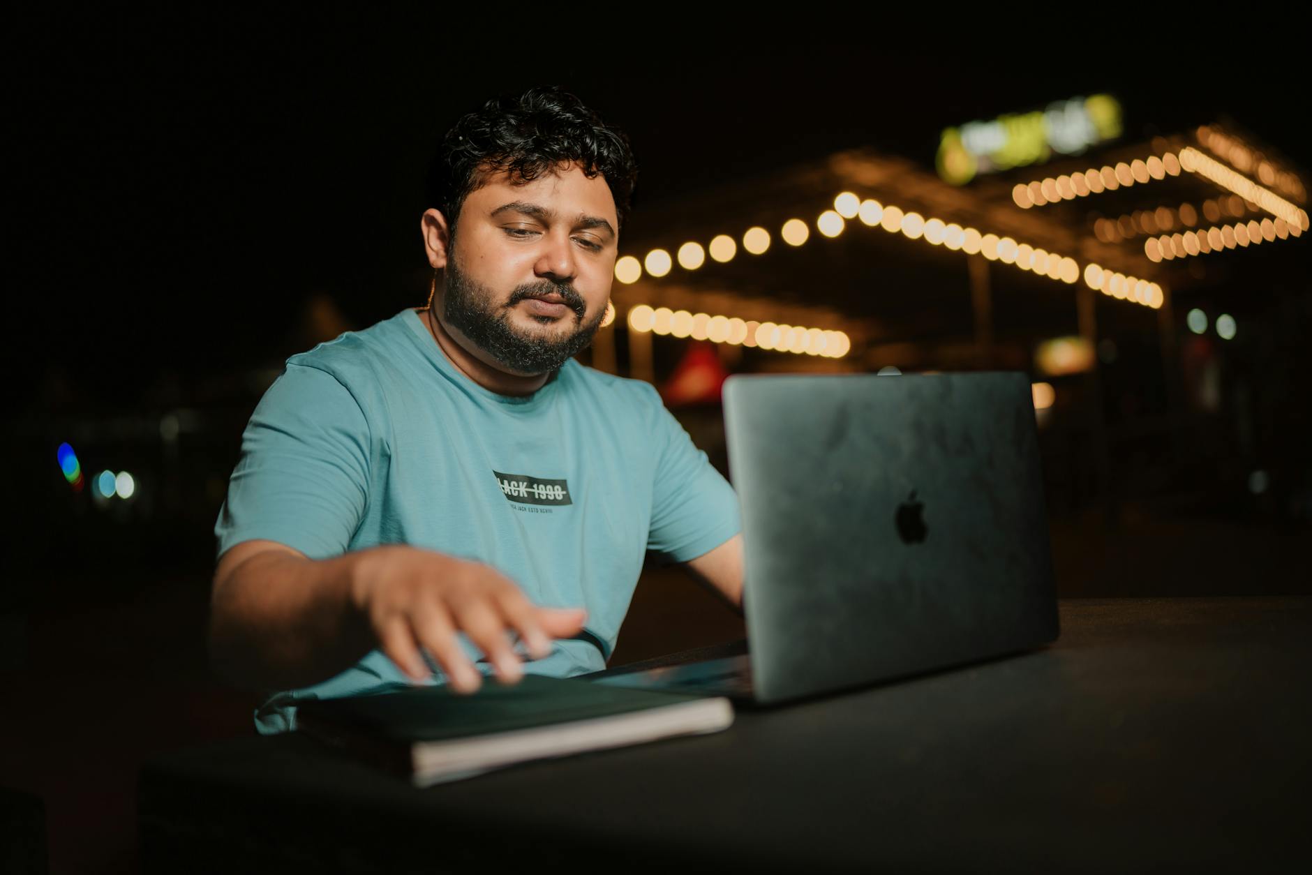 A man in a t-shirt works on a laptop at an outdoor setting at night, with decorative lights in the background.