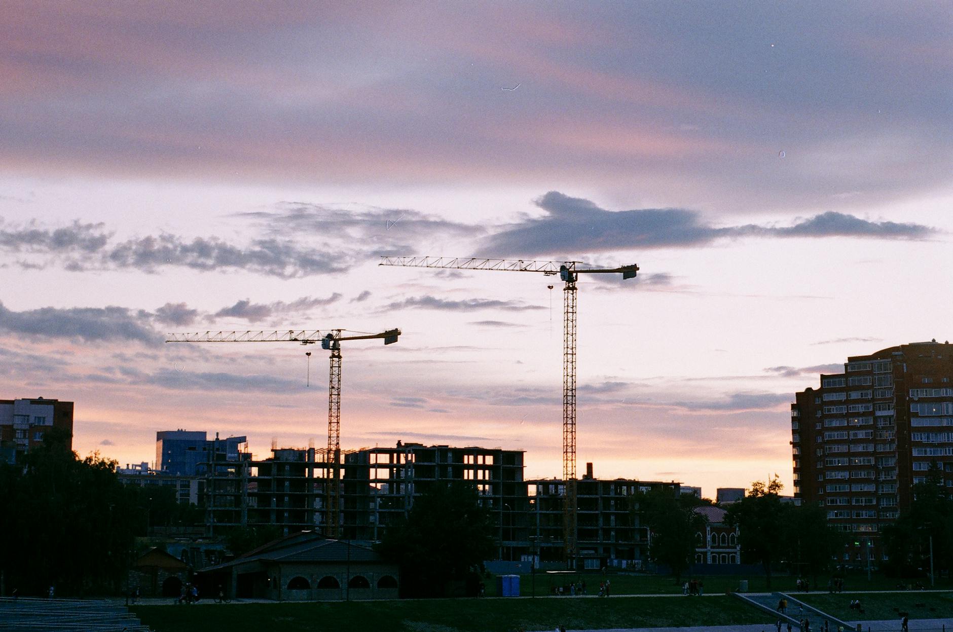 Silhouetted cranes at an urban construction site against a moody sunset sky.