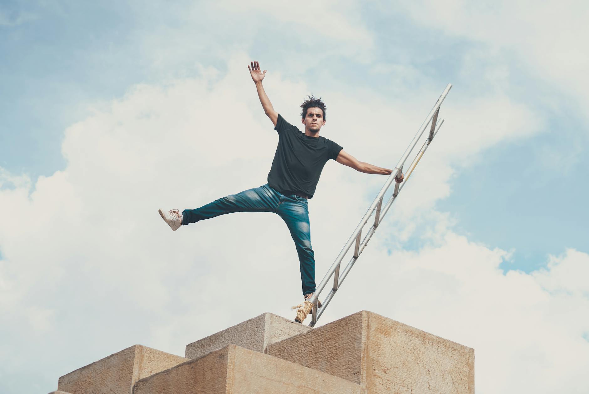 Man balancing on a ladder up high against a vibrant blue sky with clouds.