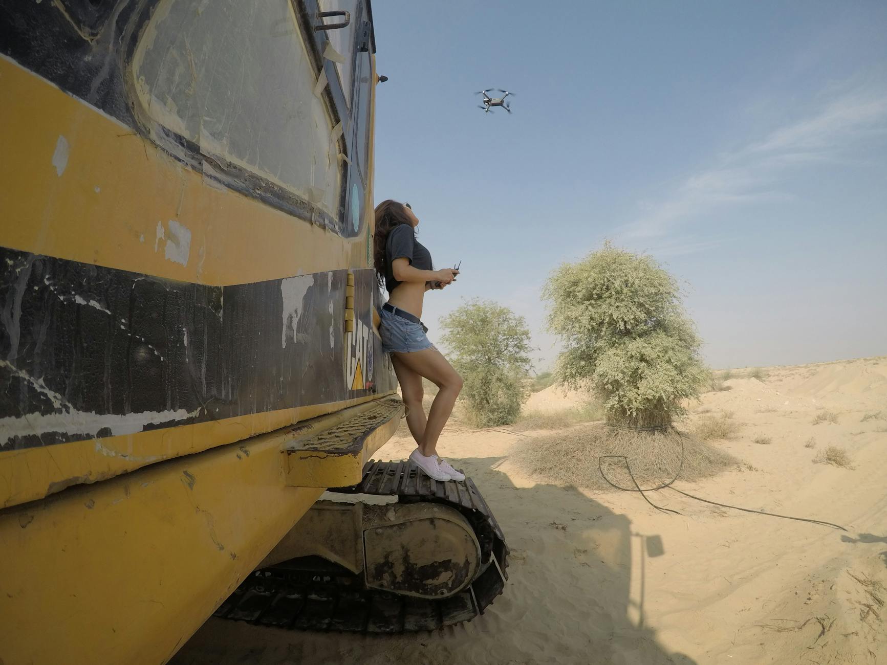 A woman controls a drone while standing on construction equipment in the Dubai desert.