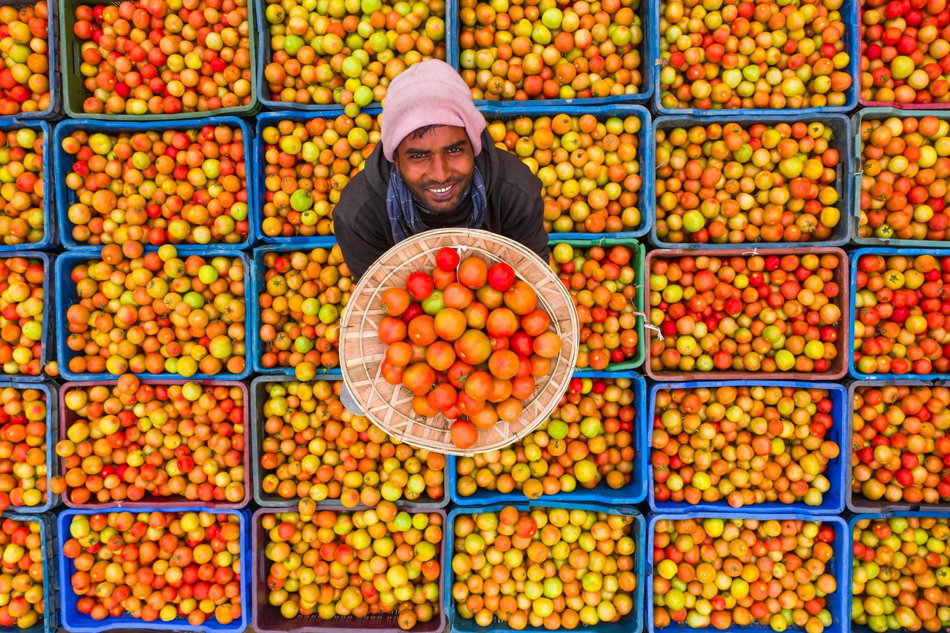 Aerial view of a smiling vendor displaying fresh tomatoes in crates at an outdoor market.