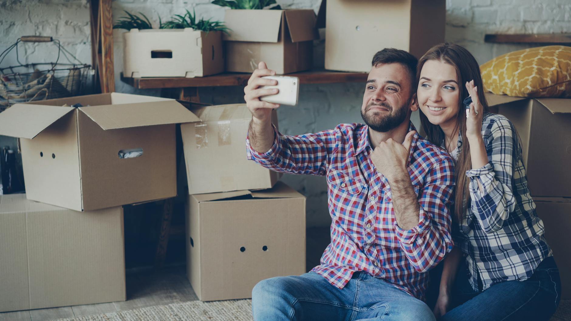 Happy couple taking a selfie while surrounded by moving boxes, celebrating new home.