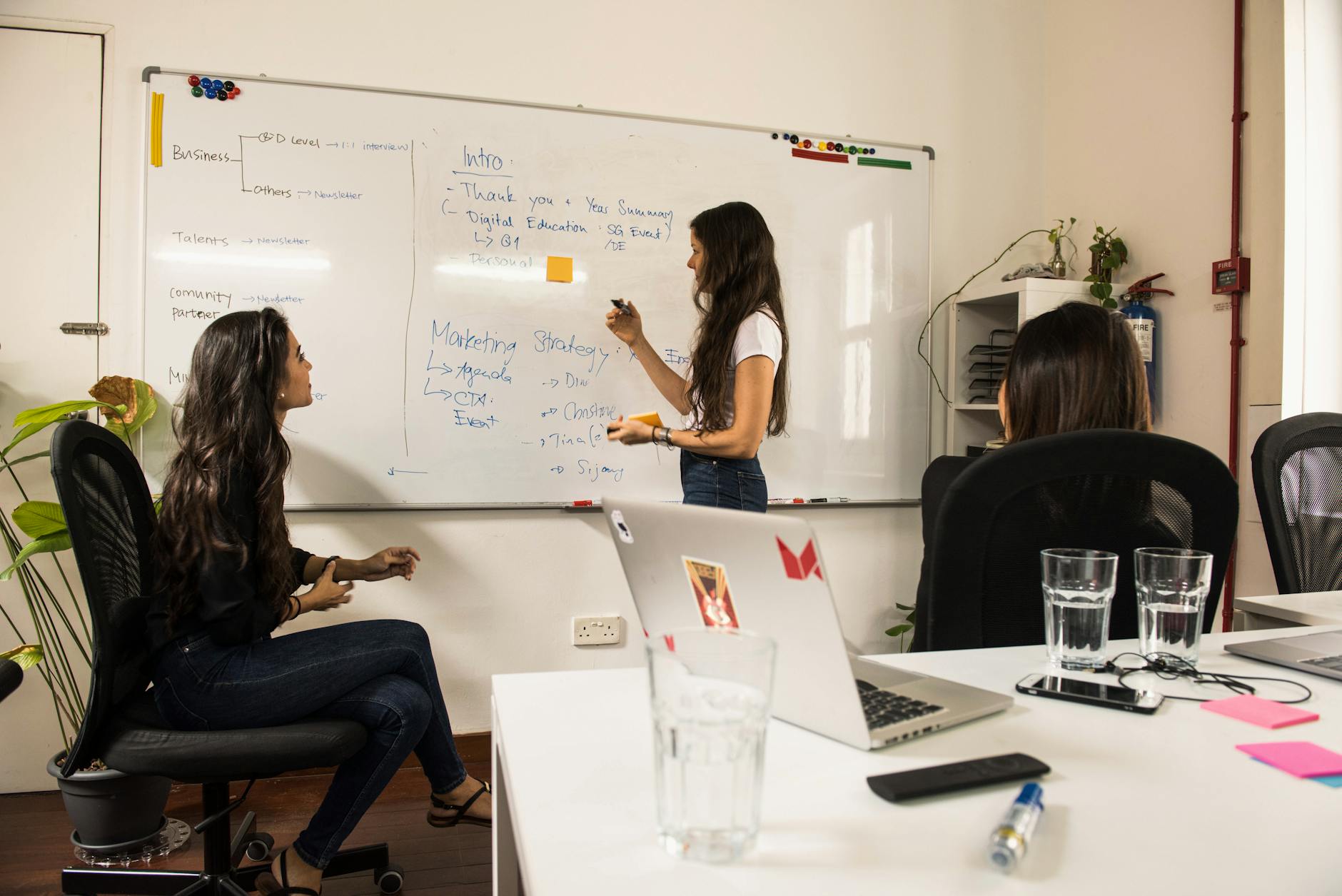 Business women discussing strategy at a whiteboard in a modern office setting.