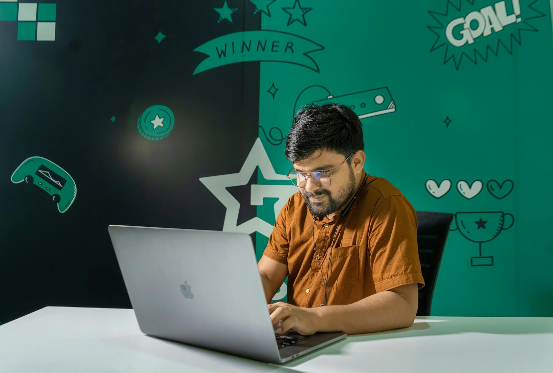 A man with a beard and glasses smiles while gaming on a laptop indoors, decorated with gaming graphics.