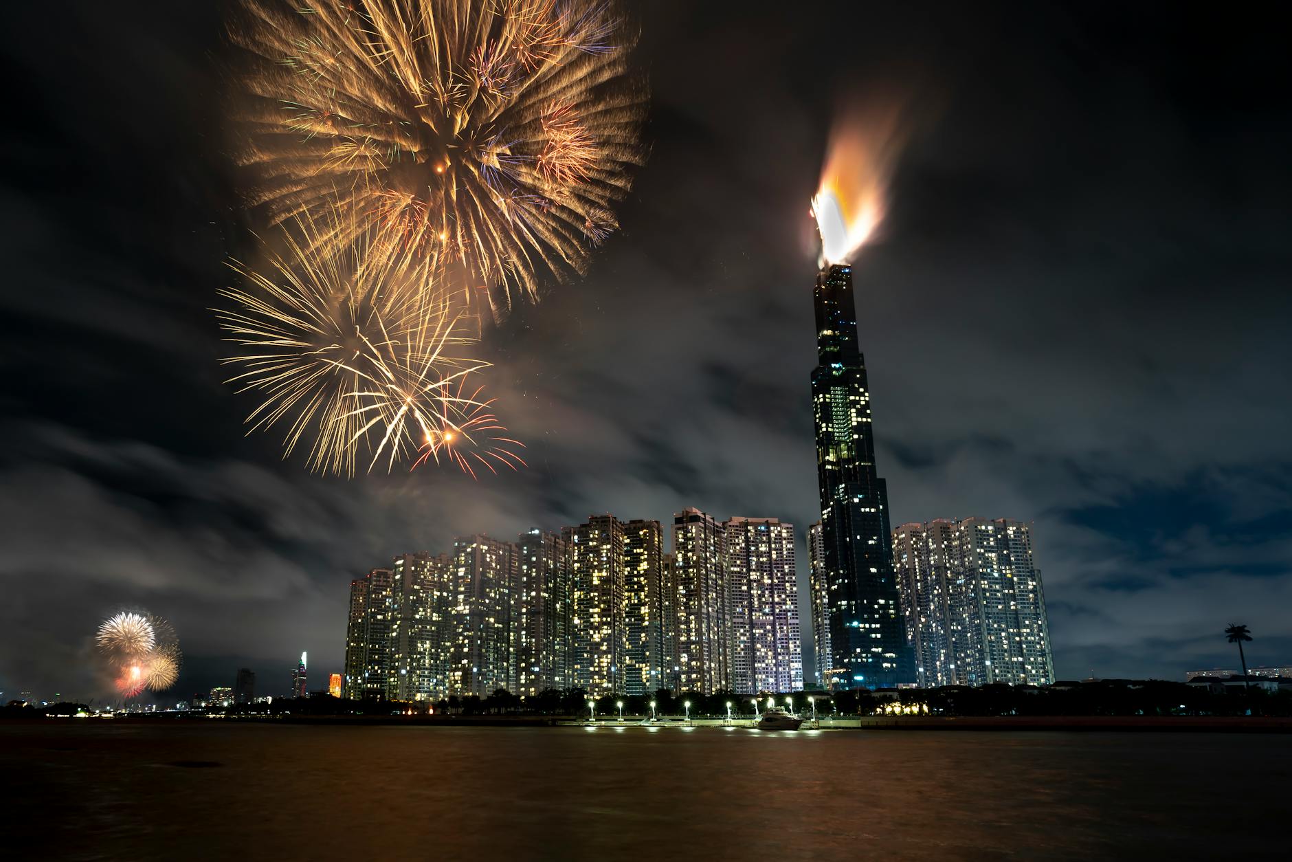 Modern glowing skyscrapers in Ho Chi Minh City in Vietnam with tall building named Landmark 81 near black water under dark cloudy sky with orange fireworks at night