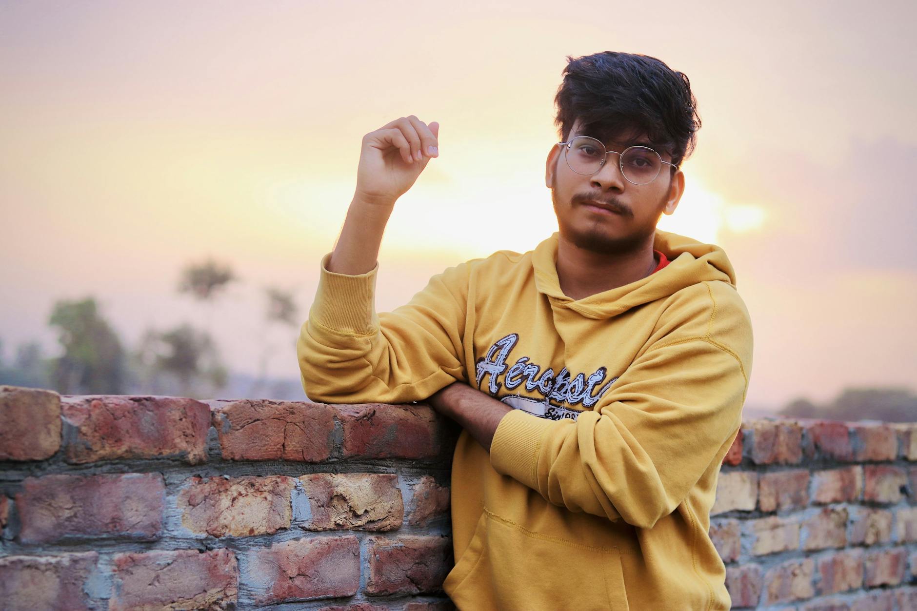 Portrait of a young man in a yellow hoodie posing casually by a brick wall at sunset.