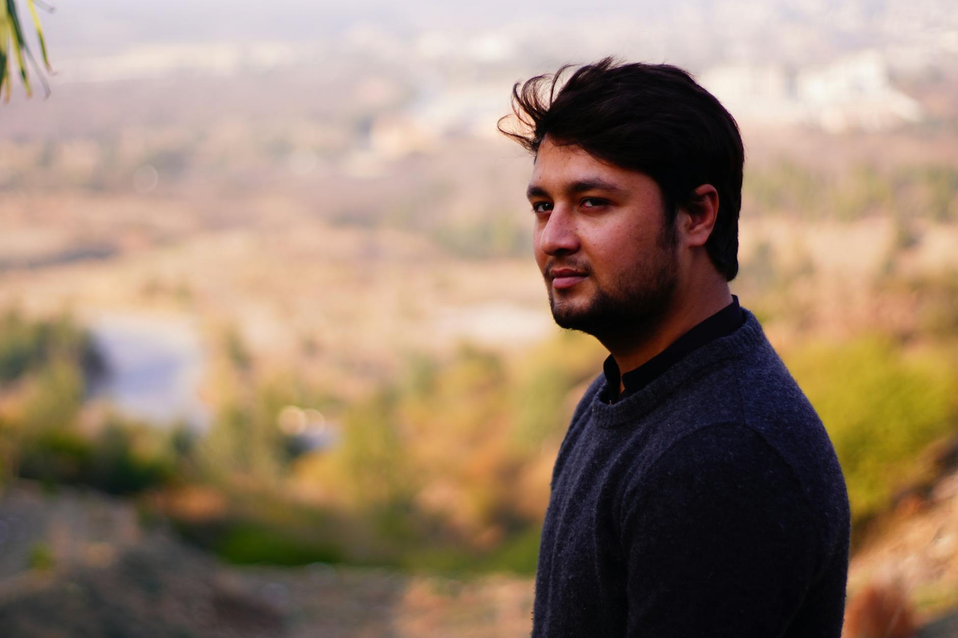A young man with a beard posing outdoors with a scenic nature backdrop.