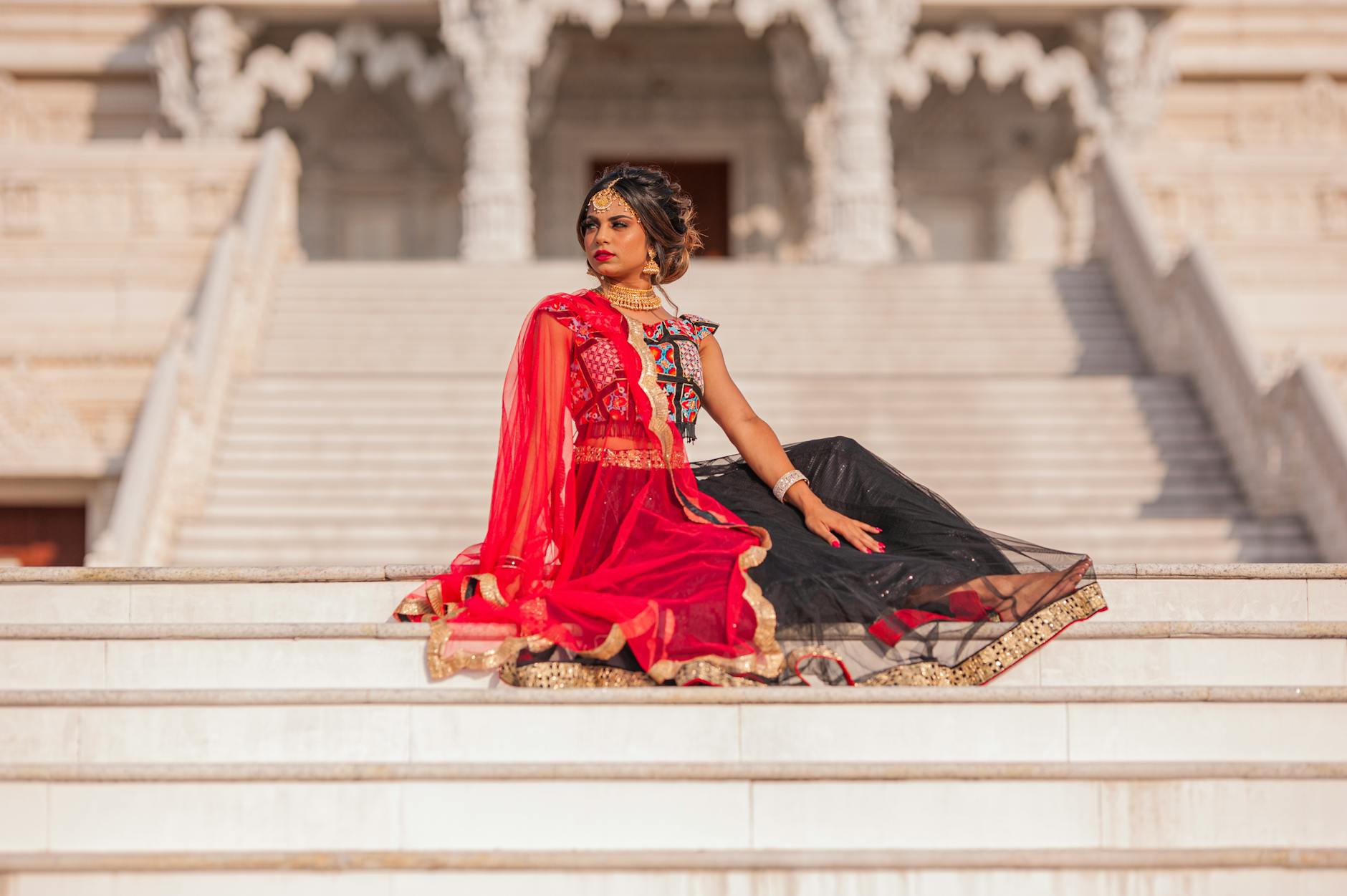 Stylish Indian woman in vibrant red and black traditional dress sitting gracefully on temple steps in Atlanta, GA.