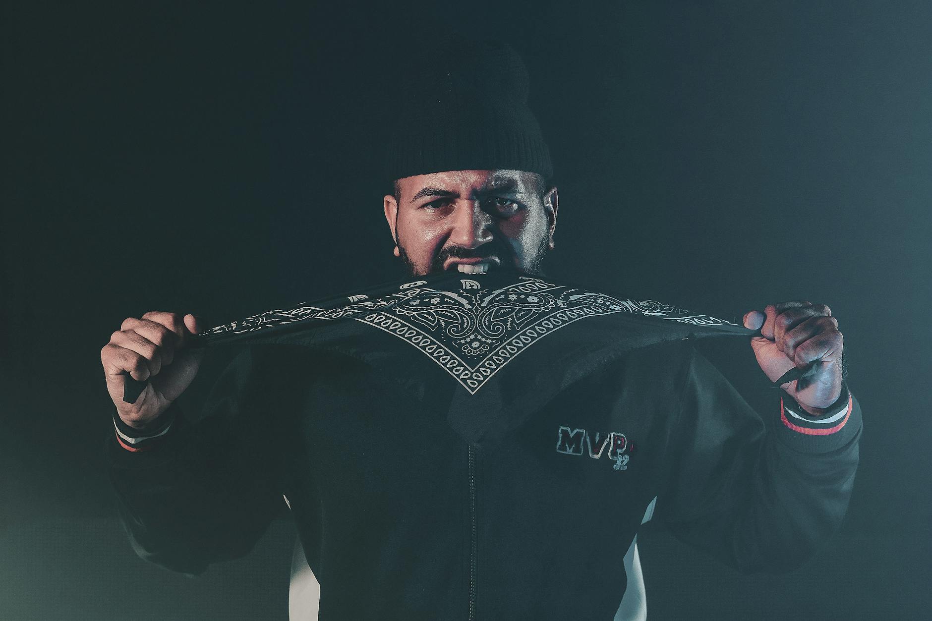 A man intensely holding a patterned bandana in a dark, moody setting.