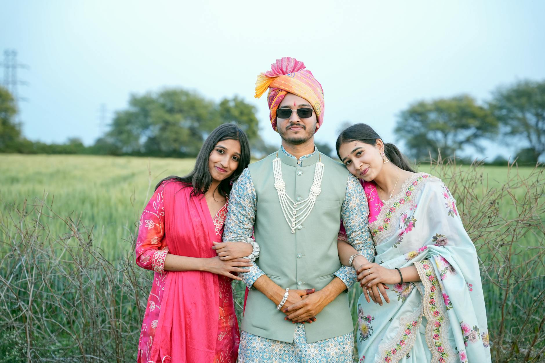 Three adults in vibrant traditional Indian attire posing in a lush green field.