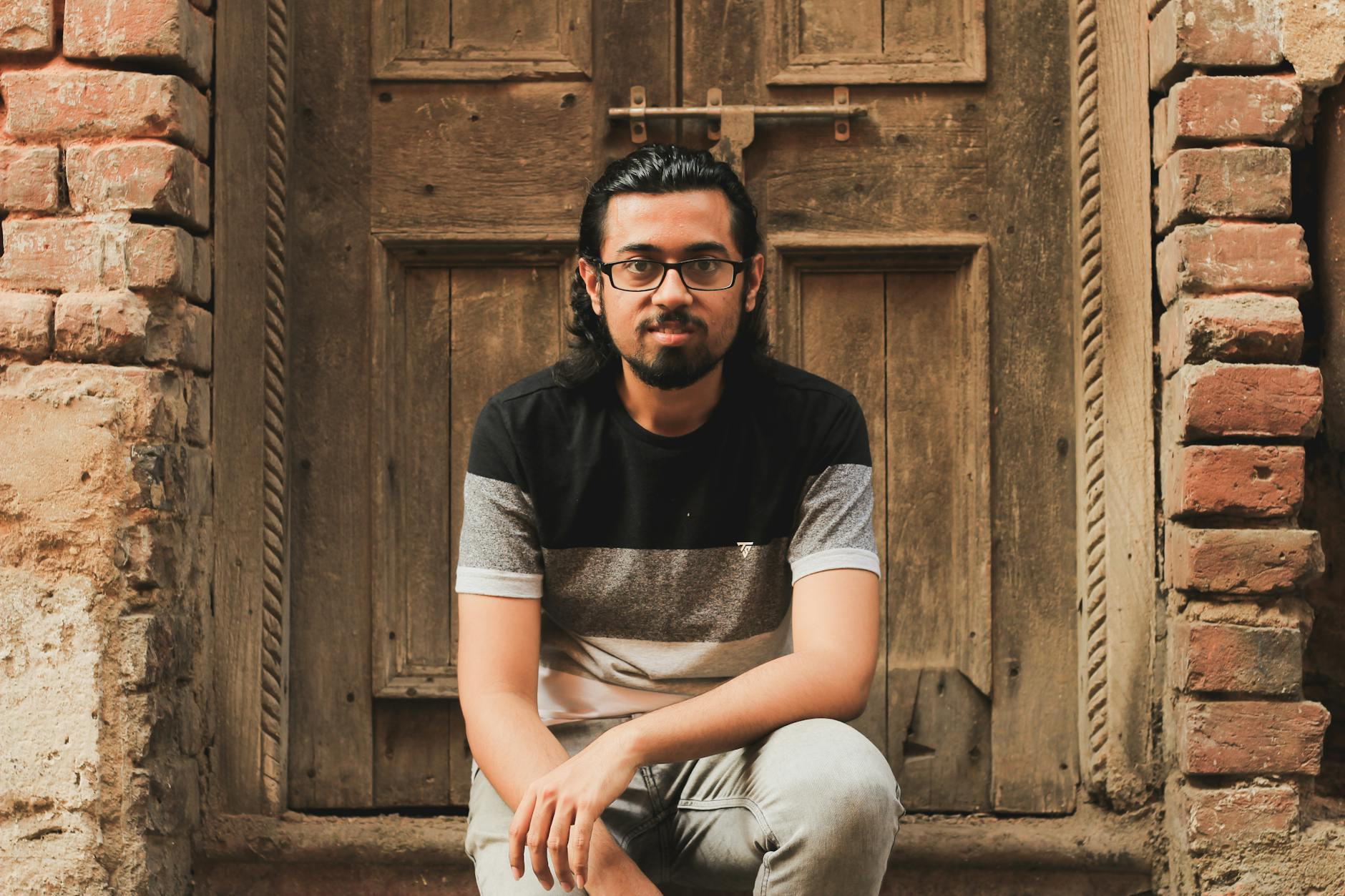 Man in casual attire sitting against an old wooden door, exuding a relaxed vibe.