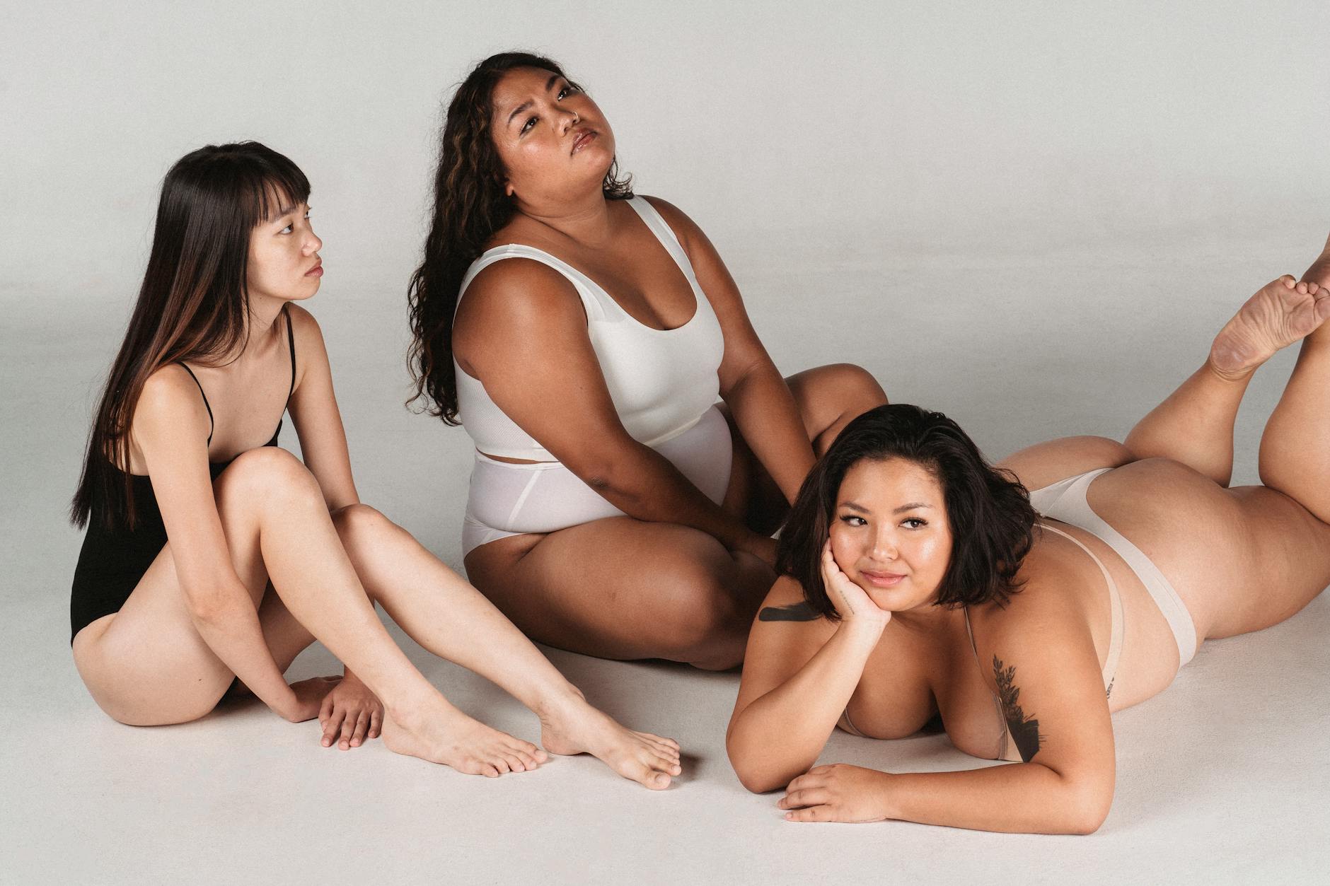 Serene Asian females with slim and plump figure types wearing lingerie resting on floor against white wall in light studio