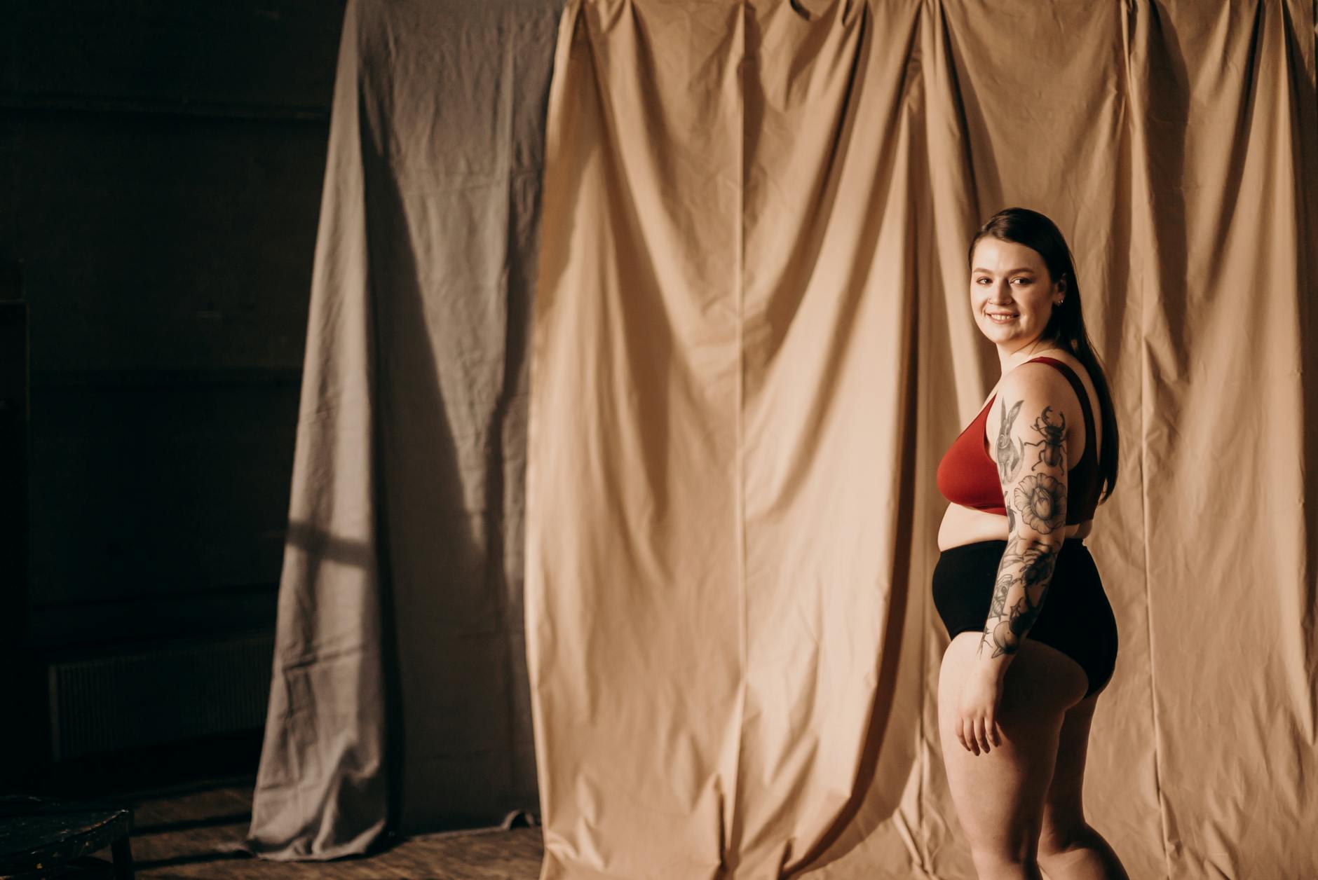 Smiling woman in lingerie with tattoo posing confidently indoors. Empowerment and body positivity theme.