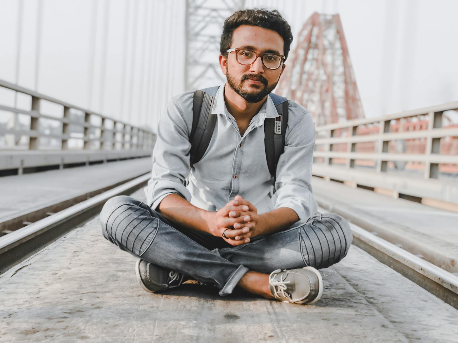 Casual portrait of a man sitting cross-legged on a bridge with an urban backdrop.
