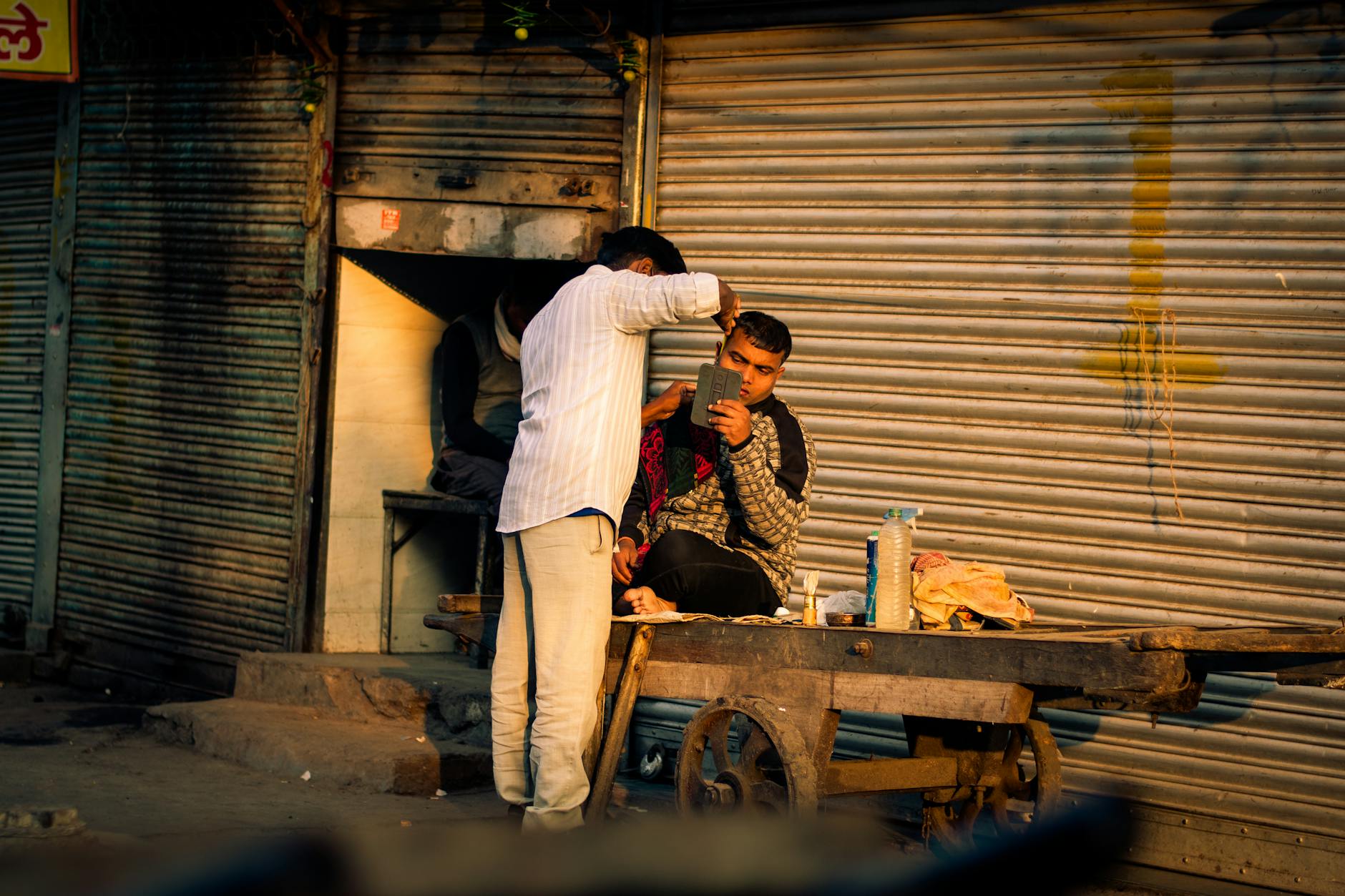 A street barber giving a haircut to a man sitting on a cart in an urban alleyway setting.
