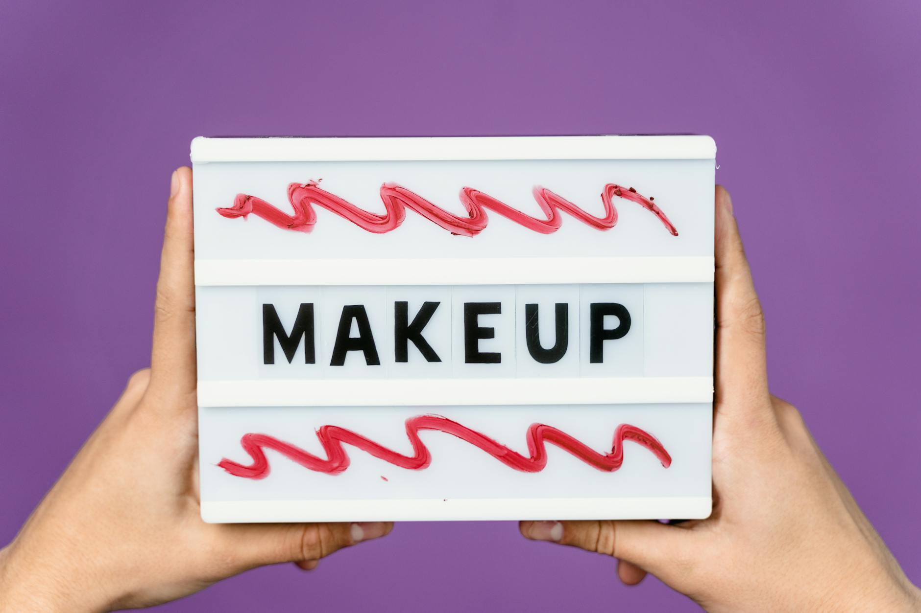 Close-up of a lightbox with the word 'MAKEUP' held by hands, featuring artistic red swirls.