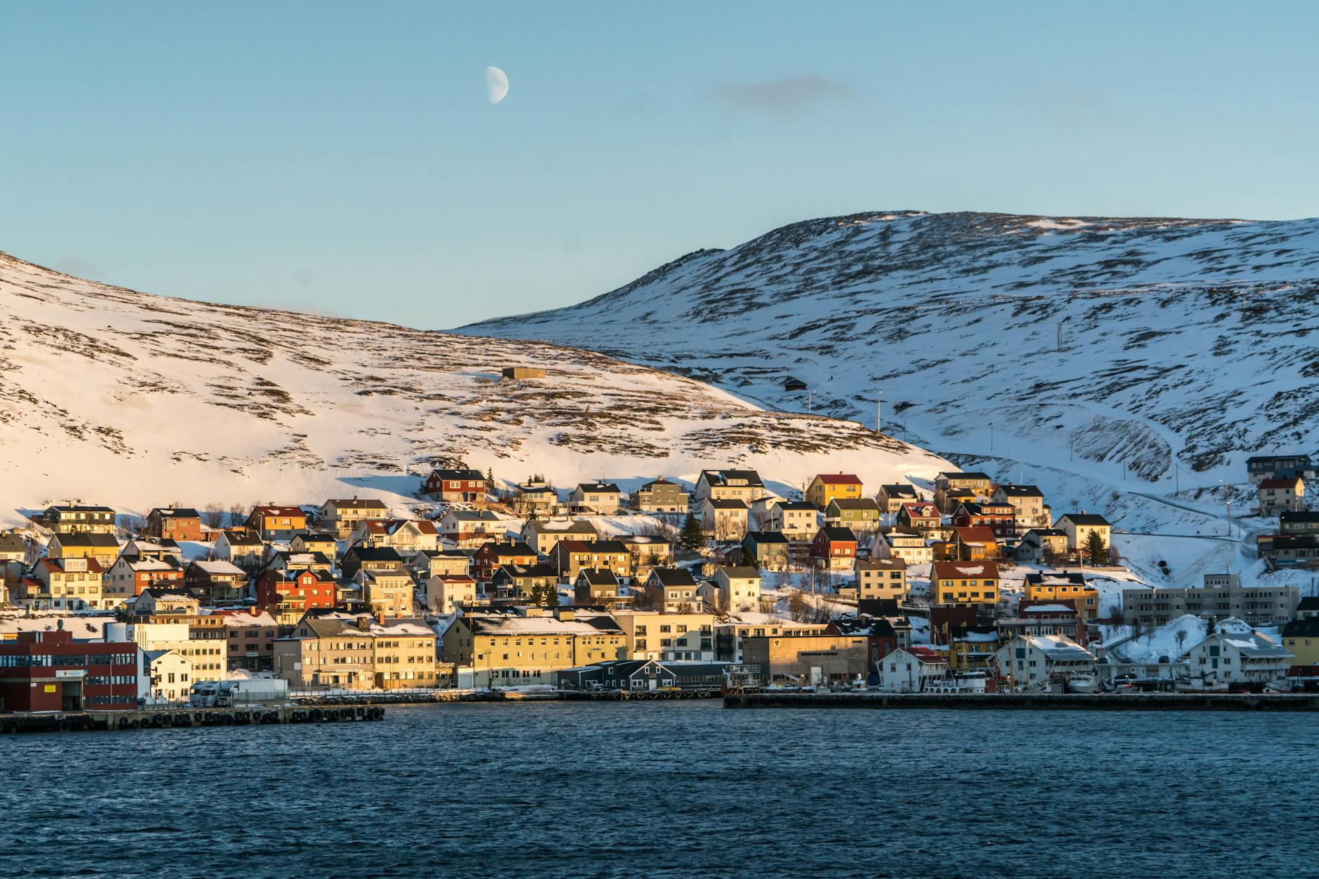 Charming Norwegian town with colorful houses on snowy hills by the sea under a bright moon.