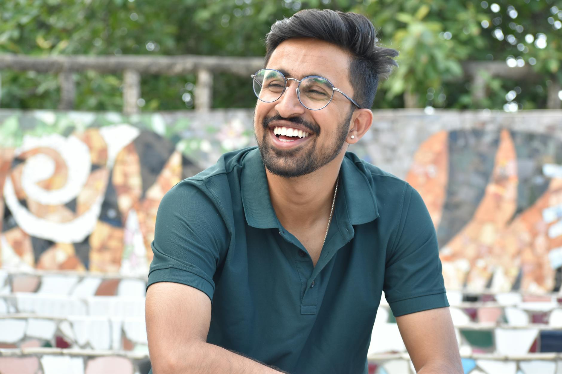 Cheerful young man with glasses smiling outdoors against artistic wall background.