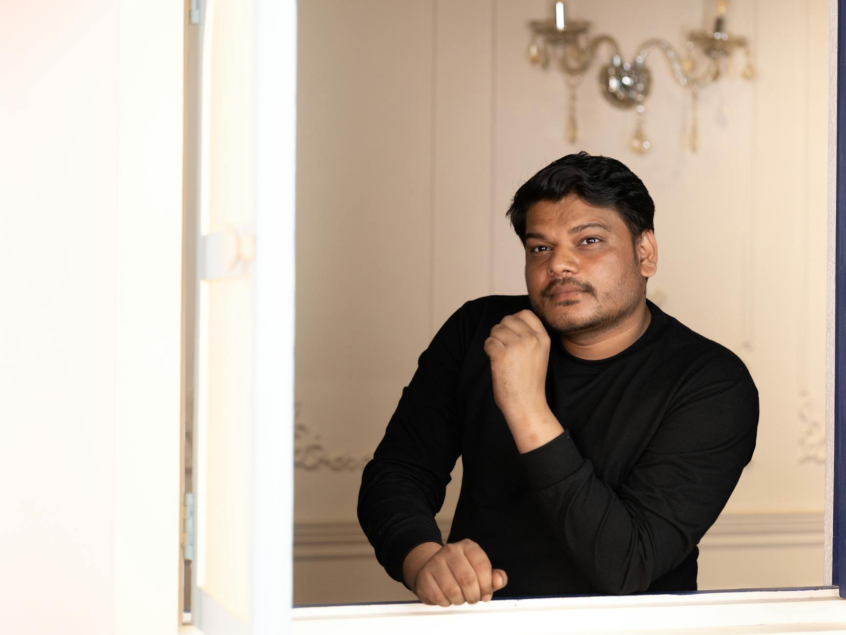 Casual portrait of a man in a black sweater leaning on a windowsill indoors.