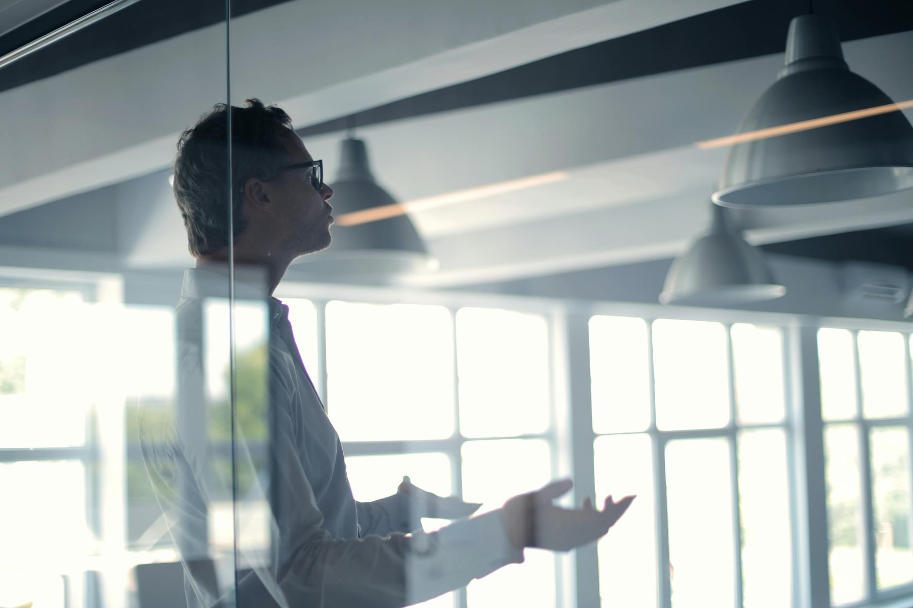 Through glass view of adult businessman in glasses and white shirt gesturing while holding presentation in spacious office