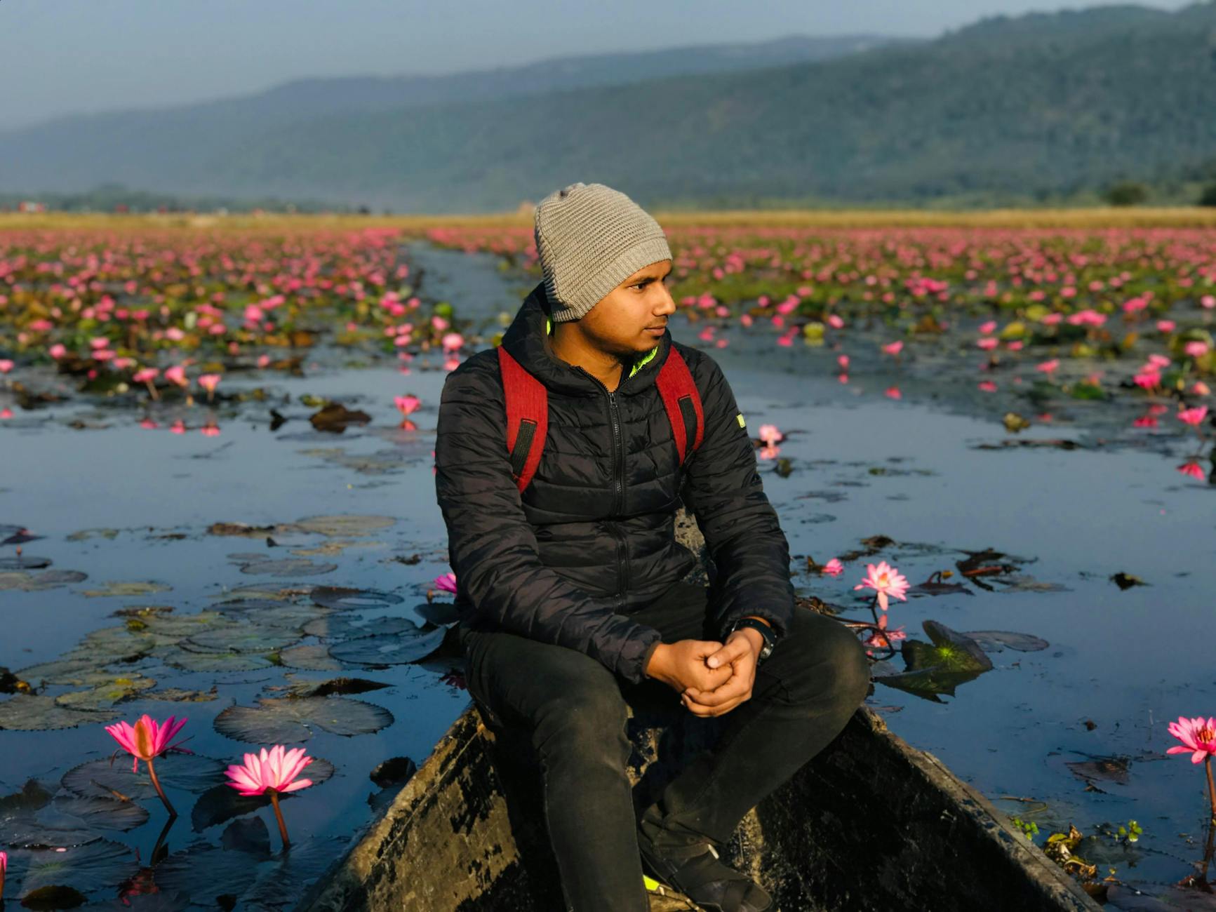 Young ethnic male traveler with backpack in knitted hat sitting on wooden boat edge while contemplating colorful flowers and leaves growing on water surface behind mounts and looking away
