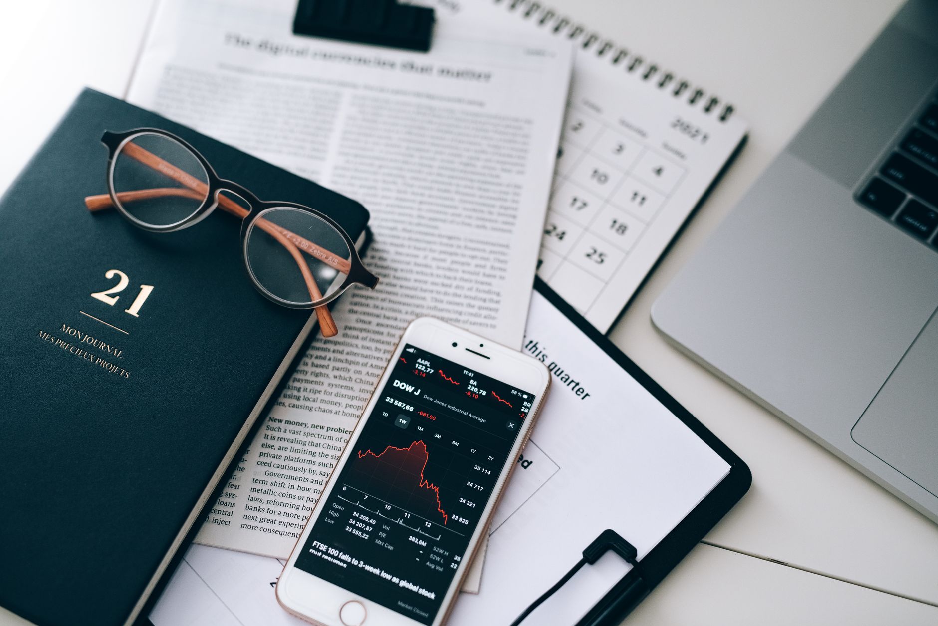 A stylish workspace featuring financial documents, eyeglasses, an iPhone displaying stock data, and a laptop.