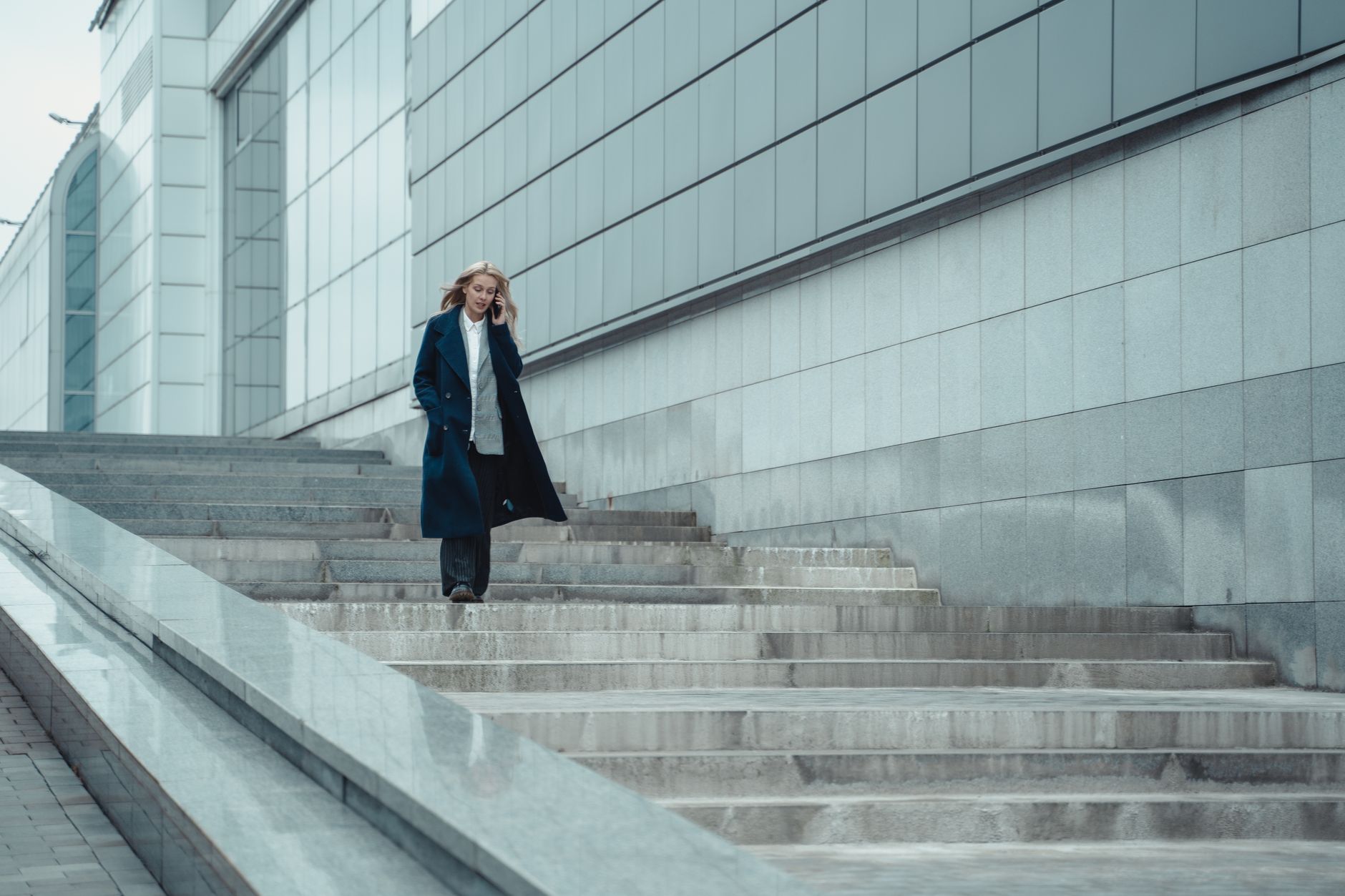 Confident woman in formal coat walking downstairs and talking on her smartphone outdoors.