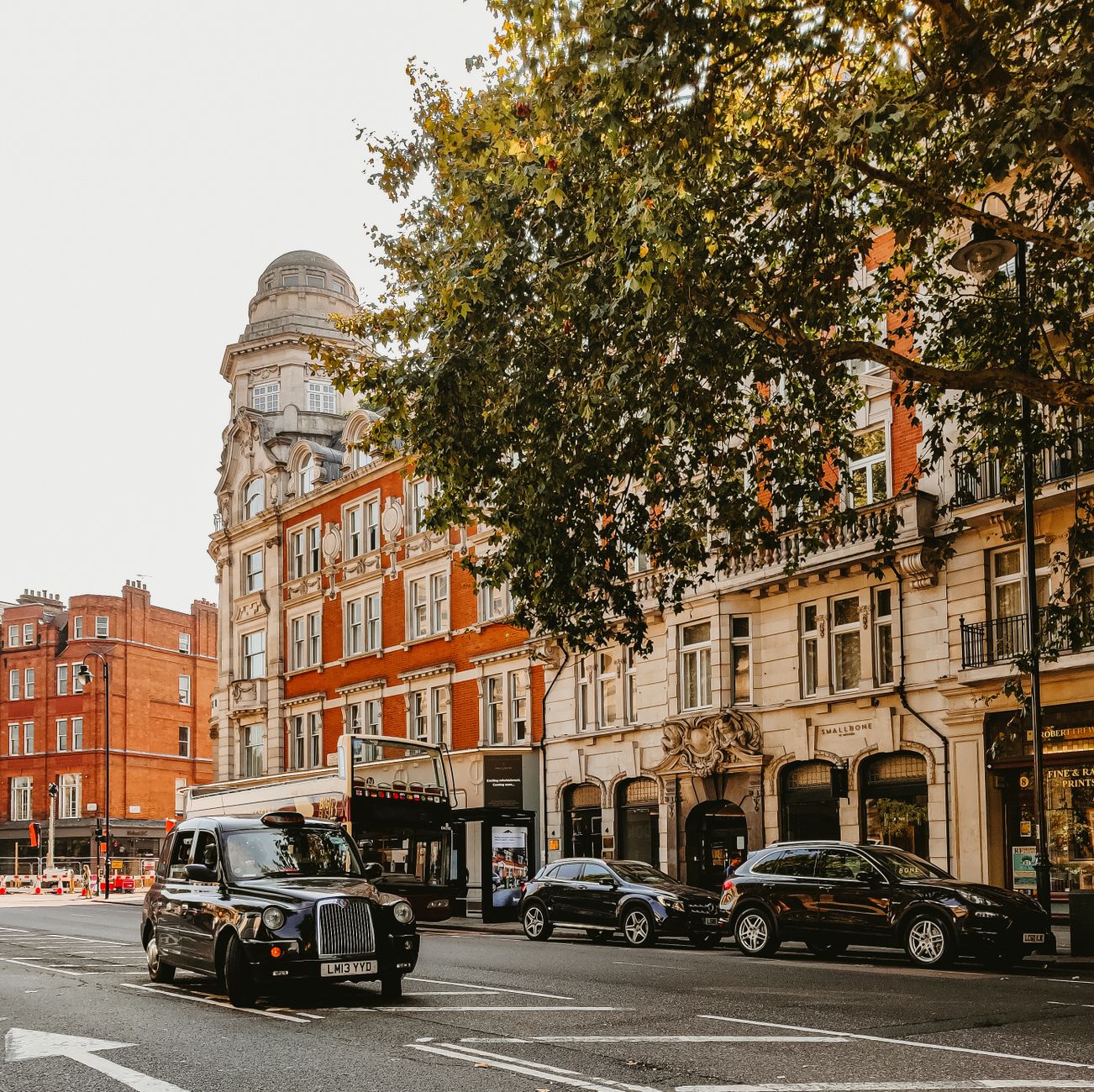 A picturesque London street featuring classic architecture and iconic black taxi under a tree's shade.